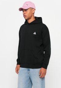 Schwarzer Kapuzenpullover mit Fronttasche und dreieckigem Logo. Getragen mit hellblauen Jeans und einer rosa Mütze. Lässiger Schnitt und schlichtes Design.