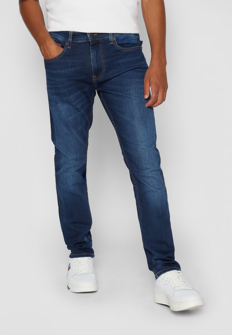 Homme portant un jean bleu foncé et des baskets blanches, debout avec une main dans la poche devant un fond uni.