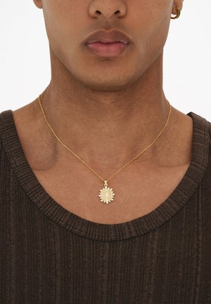 Mladá osoba nosiaca zlatý náhrdelník so slnečným náboženským príveskom a kruhovou náušnicou, oblečená v hnedom rebrovanom tričku.