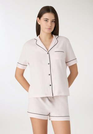 Giovane donna che indossa un pigiama rosa chiaro con bottoni a maniche corte e pantaloncini abbinati con rifiniture nere, in piedi davanti a uno sfondo bianco.