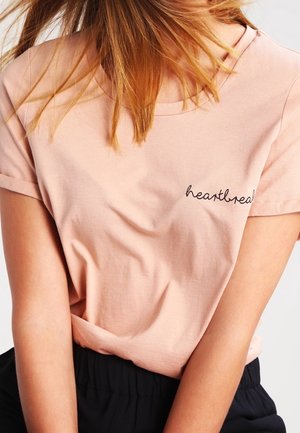 Kobieta nosząca jasnoróżowy t-shirt z wyszywanym napisem „heartbreak” w okolicy klatki piersiowej, w połączeniu z czarnymi spodniami. Włosy częściowo zasłaniają twarz.