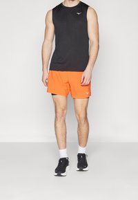 Černý bezrukávový sportovní top a oranžové šortky. Hladká tkanina, moderní střih. Černé sportovní boty s bílými podrážkami a bílé ponožky.