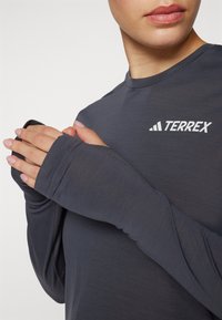 Graues Langarmshirt aus glattem Stoff mit genähten Nähten, ausgestattet mit einem Daumenloch-Design und einem weißen TERREX-Logo auf der Brust.