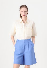 Camicia in lino crema a maniche corte con due tasche frontali, abbinata a pantaloni corti blu a vita alta con pieghe, in piedi davanti a uno sfondo bianco.