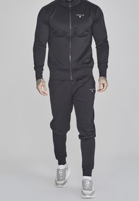 Chándal deportivo negro hecho de una tela suave, con chaqueta con cremallera y pantalones ajustados, que presenta un pequeño logo en el pecho y el muslo.