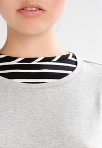 Close-up van het onderste deel van iemands gezicht en nek, met een grijze top over een zwart-wit gestreept shirt, tegen een effen lichte achtergrond.