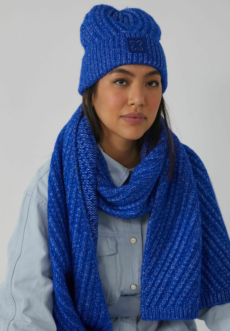 Blauwe gebreide muts met een gestructureerd patroon en een logo patch, gecombineerd met een bijpassende geribbelde sjaal. Beide tonen een levendige tint blauw.