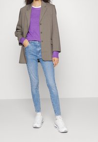 Blazer gris sobre un suéter púrpura, combinado con jeans ajustados de color azul claro y zapatillas blancas. Las características destacadas incluyen un diseño entallado y detalles de botones.