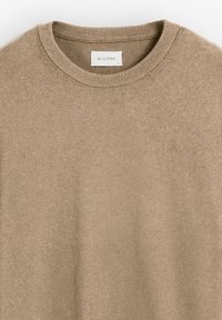 Suéter de punto beige con un escote redondeado y borde acanalado. Presenta un sutil logo bordado en el pecho. Textura suave y diseño liviano.