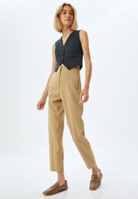 Estriiped navy cropped vest dengan leher v, dipadukan dengan celana panjang beige high-waisted. Model mengenakan sepatu mokasi berbahan tekstur coklat, berdiri di latar belakang putih.