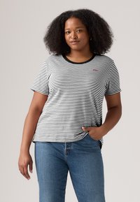 Kvinne med krøllete hår iført svart-hvitt stripete T-skjorte og blå jeans, stående med den ene hånden i lommen mot grå bakgrunn.