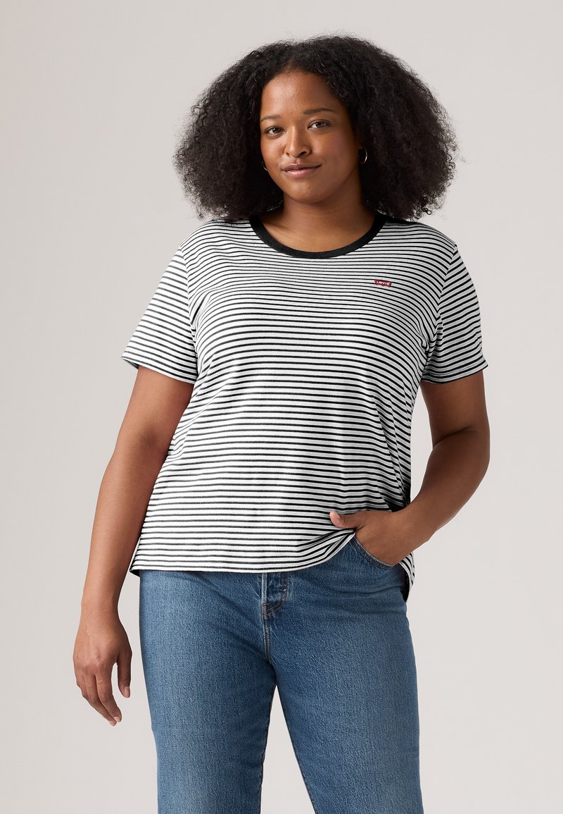 Kvinne med krøllete hår iført svart-hvitt stripete T-skjorte og blå jeans, stående med den ene hånden i lommen mot grå bakgrunn.