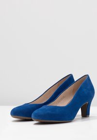 Pompes à talons hauts en suede bleu avec des bouts ronds, une surface légèrement texturée et un talon robuste. L'intérieur est doublé de couleur chair.