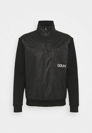 Maglione nero con mezza zip frontale, collo alto, maniche lunghe, ampie tasche sul petto a sinistra con logo "HUGO" bianco capovolto.