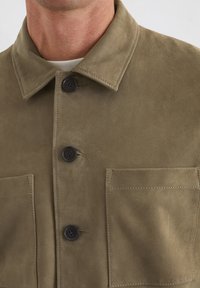 Wildlederjacke in Olivgrün mit einem Kragen, zwei Fronttaschen und schwarzen Knöpfen. Glatte Textur mit sichtbaren Nähdetails.