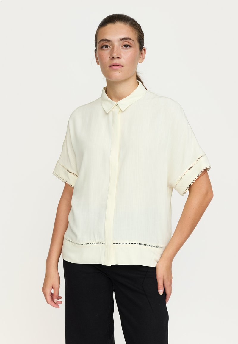 Femme aux cheveux bruns en queue de cheval basse porte une chemise blanche à manches courtes boutonnée et un pantalon noir, debout devant un fond clair uni.