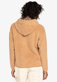 Tan fleece hoodie with a hood, showing a soft texture, relaxed fit, and a clean, simple design. The garment has a slightly cropped cut at the waist.