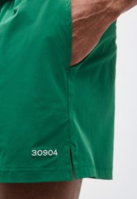 Χέρι στην τσέπη πράσινου σόρτς με μικρή πλευρική σχισμή και λευκό κεντημένο αριθμό "30904" κοντά στο τελείωμα.