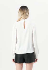 White long-sleeve blouse with puffed sleeves and a back keyhole detail, paired with black leather shorts. Smooth texture, casual design.