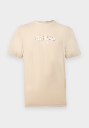 Tricou bej cu mâneci scurte, cu logo-ul alb „GUESS EST. 1981” centrat pe piept, pe un fundal simplu.