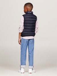 Gilet puffer blu navy con colletto decorato con un logo, abbinato a una camicia a righe chiara, jeans azzurri arrotolati e sneakers bianche.
