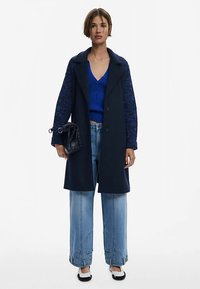 Tmavě modrý kabát z vlněné směsi s texturovanými rukávy, nošený přes modrý žebrovaný top. Modelka má na sobě světle modré široké džíny a nese černou tašku.