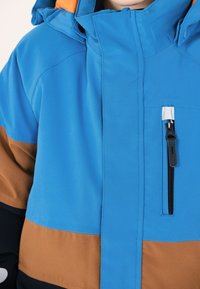 Blå och brun färgblockerad jacka med hög krage, blixtlåsförsedd bröstficka, och orangea detaljer på huven. Slät tygerstruktur.