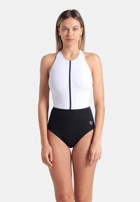 Maillot de bain à col montant avec un haut blanc et un bas taille haute noir. Présente une fermeture éclair noire centrale et un détail de logo sur la hanche gauche.