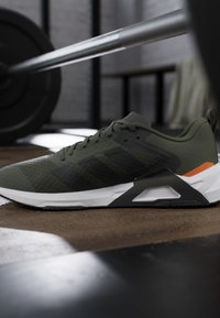 Chaussure de sport verte et noire avec semelle blanche sur le sol de la salle de sport près d'une barre avec des poids dans un cadre d'entraînement fitness.