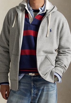 Homme portant un sweat à capuche gris clair zippé, un maillot de rugby rayé bleu marine et rouge par-dessus une chemise bleue à boutons, un jean bleu et une montre-bracelet.