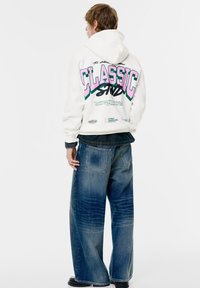 Witte hoodie met "CLASSIC" in roze en groen op de achterkant, gecombineerd met wijde, verweerde blauwe jeans. Opvallende borduur- en grafische details.