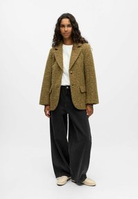 Bruine, gestructureerde wollen blazer met piekflappen, twee knopen en voorzakken, gecombineerd met loszittende zwarte denim broeken en lichte schoenen.