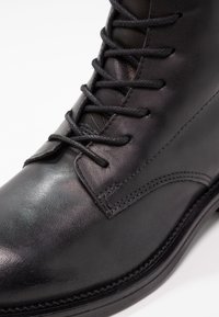 Bottine en cuir noir avec des lacets œillet, texture lisse et détails de couture surélevés le long de la semelle et du capuchon de orteil.