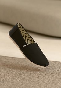 Sorte lerret slip-on sko med rund tå og hvit gummisåle. Innvendig har den et sikksakkmønster i beige og mørkebrunt.