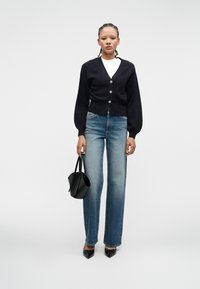 Woman wearing a black cardigan, white shirt, blue jeans, and black pointed shoes, holding a black handbag against a plain white background.