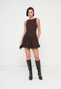 Καφέ αμάνικο φόρεμα με εφαρμοστό τοπ και φουσκωτή φούστα με λεπτομέρειες κορδονιών. Συνδυάζεται με μαύρες δερμάτινες μπότες έως το γόνατο.