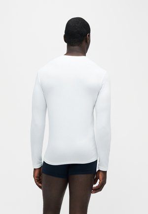 Mann mit dunkler Haut, der ein schlichtes weißes Langarmshirt und dunkle Boxershorts trägt, mit dem Rücken zur Kamera auf weißem Hintergrund.