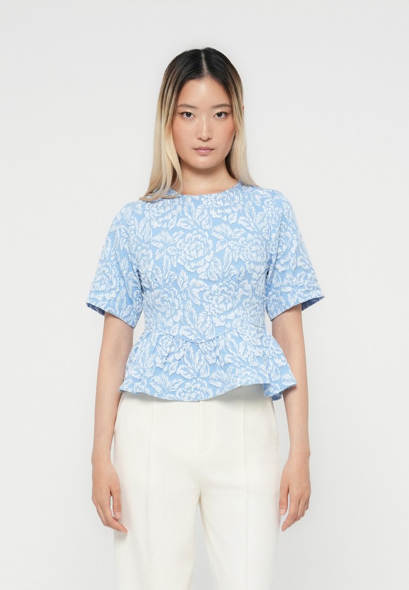 Hellblaues Peplum-Top mit weißem Blumenmuster, kurzen Ärmeln und rundem Ausschnitt. Weicher Stoff mit glatter Textur. Kombiniert mit weißen Hosen.