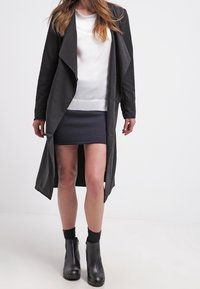 Svart lång kappa med draperad krage, över en vit blus och texturerad marinblå kjol, kombinerad med svarta ankelstövlar. Släthet, modern design.