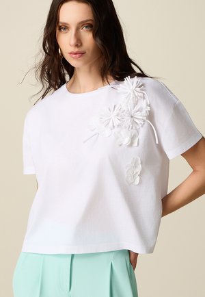 Donna con una maglietta bianca a maniche corte con applicazioni floreali 3D sulla spalla e pantaloni verde menta a vita alta, in piedi con una mano sul fianco.