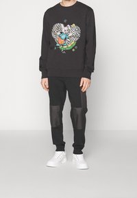 Felpa nera con una grafica cartoon a forma di cuore, abbinata a pantaloni jogger neri con pannelli trasparenti. Completata con sneaker bianche.