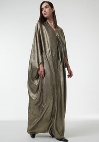Robe longue métallisée dorée avec un décolleté en V, des manches amples et un tissu drapé. Présente une texture lisse et un design long jusqu'aux chevilles, accompagnée de talons noirs.