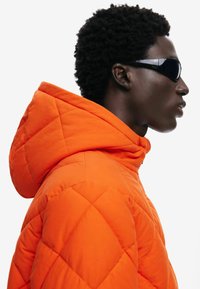 Giacca puffer arancione trapuntata con colletto alto e cappuccio integrato; il modello indossa occhiali da sole neri. Superficie texture con cuciture visibili.