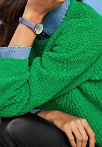 Persoon draagt een groene gebreide trui en een spijkerbloes met een lavendelkleurig horloge aan de linkerpols, hand rust tegen het gezicht voor een blauwe achtergrond.