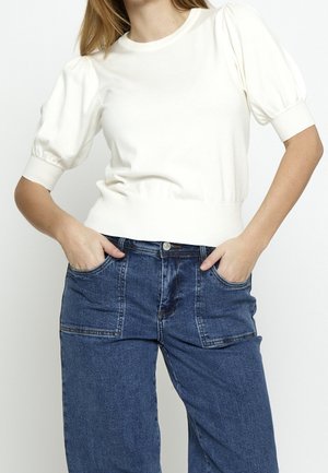 Femme portant un haut en maille blanc à manches bouffantes et un jean en denim bicolore taille haute, les mains dans les poches avant sur fond blanc.