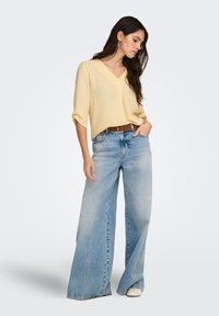 Geel, top met korte mouwen en V-hals, gecombineerd met wijd uitlopende lichtblauwe jeans. Bruine riem benadrukt de taille. Licht materiaal en losse pasvorm.