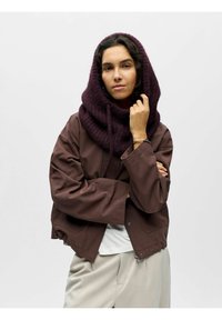 Veste marron coupée courte avec des manches amples, associée à une capuche en tricot violet foncé. Le mannequin porte un haut blanc, présentant un look neutre.