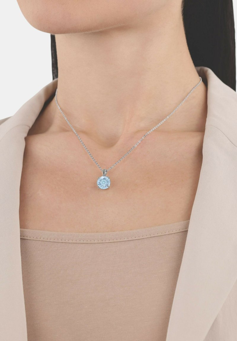 Silverkedja med en rund, ljusblå ädelsten som hängs över en beige topp och under en ljusbeige blazer.