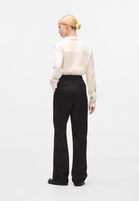 Blusa bianca a maniche lunghe con vestibilità leggermente rilassata e yoke sul retro, abbinata a pantaloni neri a vita alta con motivo e scarpe nere.