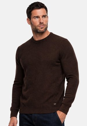 Uomo con capelli corti e scuri e barba, che indossa un maglione marrone scuro a maniche lunghe e jeans blu scuro con una mano in tasca.